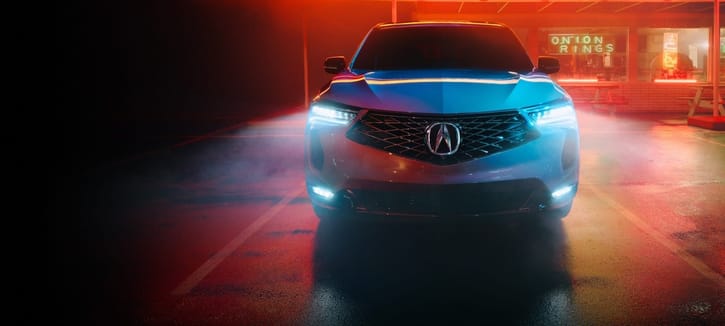 A sleek 2025 Acura RDX is illuminated in neon lights, with a foggy parking lot backdrop showcasing a glowing onion rings sign.
