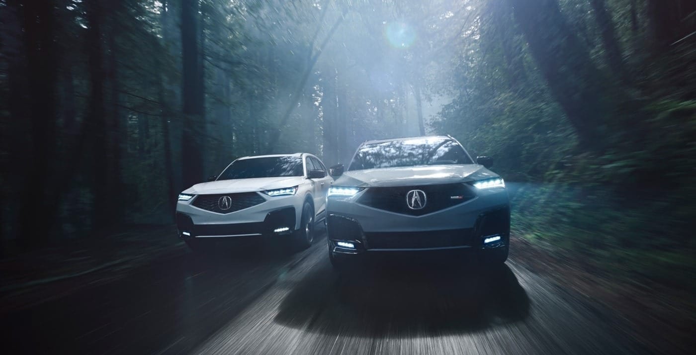Two 2026 Acura MDX SUVs, one white and one gray, drive through a misty forest, showcasing their sleek designs under soft, diffused light.