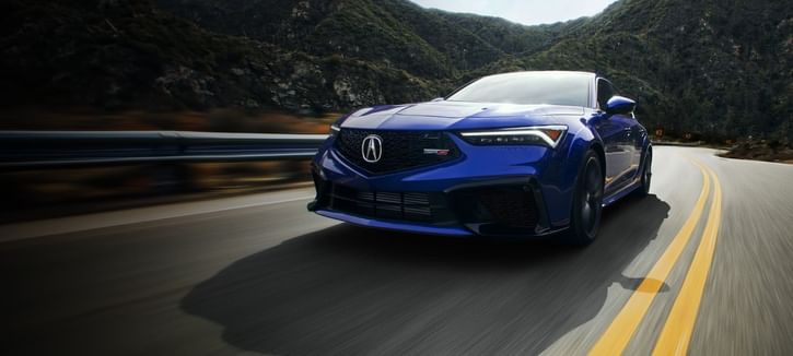 A blue 2025 Acura Integra driving at high speed on a winding road, surrounded by hills and greenery under a bright sky.