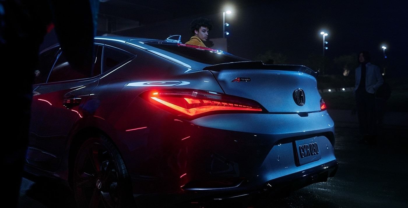 A sleek silver 2025 Acura Integra with aggressive styling and illuminated taillights, parked under dim lights in a nighttime setting.