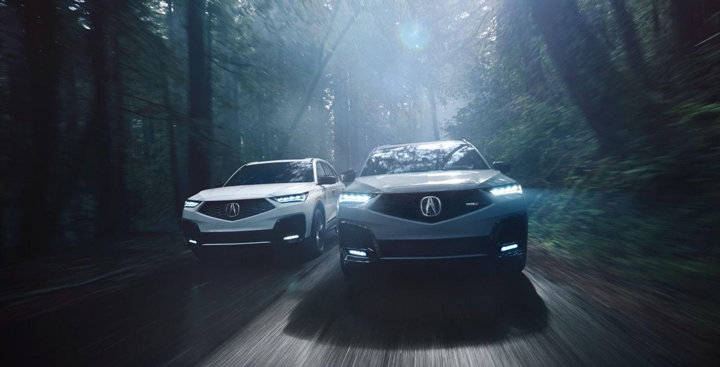 Two 2026 Acura MDX SUVs, one white and one gray, drive through a misty forest, showcasing their sleek designs under soft, diffused light.