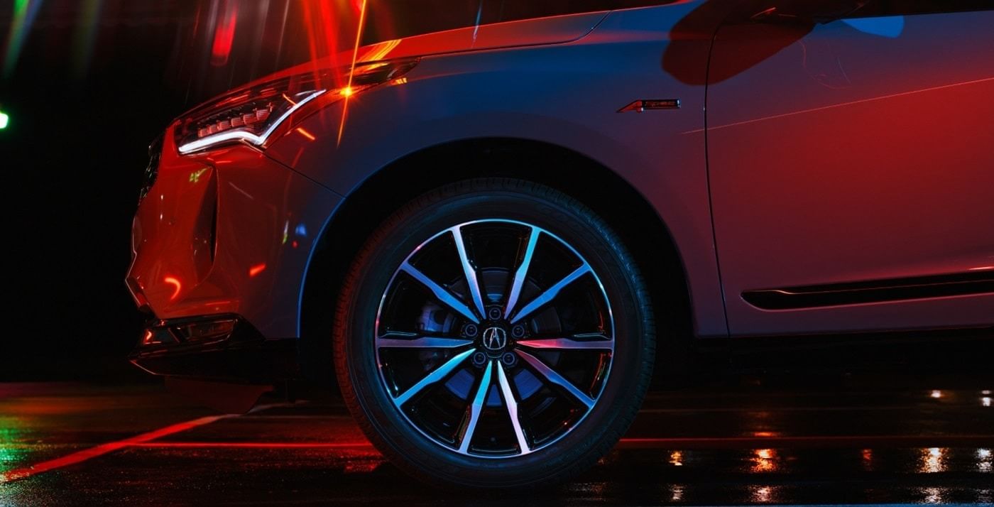 Close-up of a 2025 Acura RDX wheel with glossy black alloy rims, illuminated by colorful lights on a wet surface.