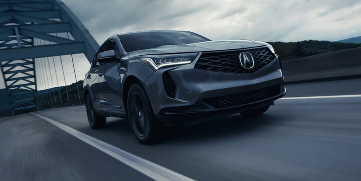A dark gray 2026 Acura RDX driving dynamically on a bridge under an overcast sky, highlighting its diamond pentagon grille and sharp LED headlights.