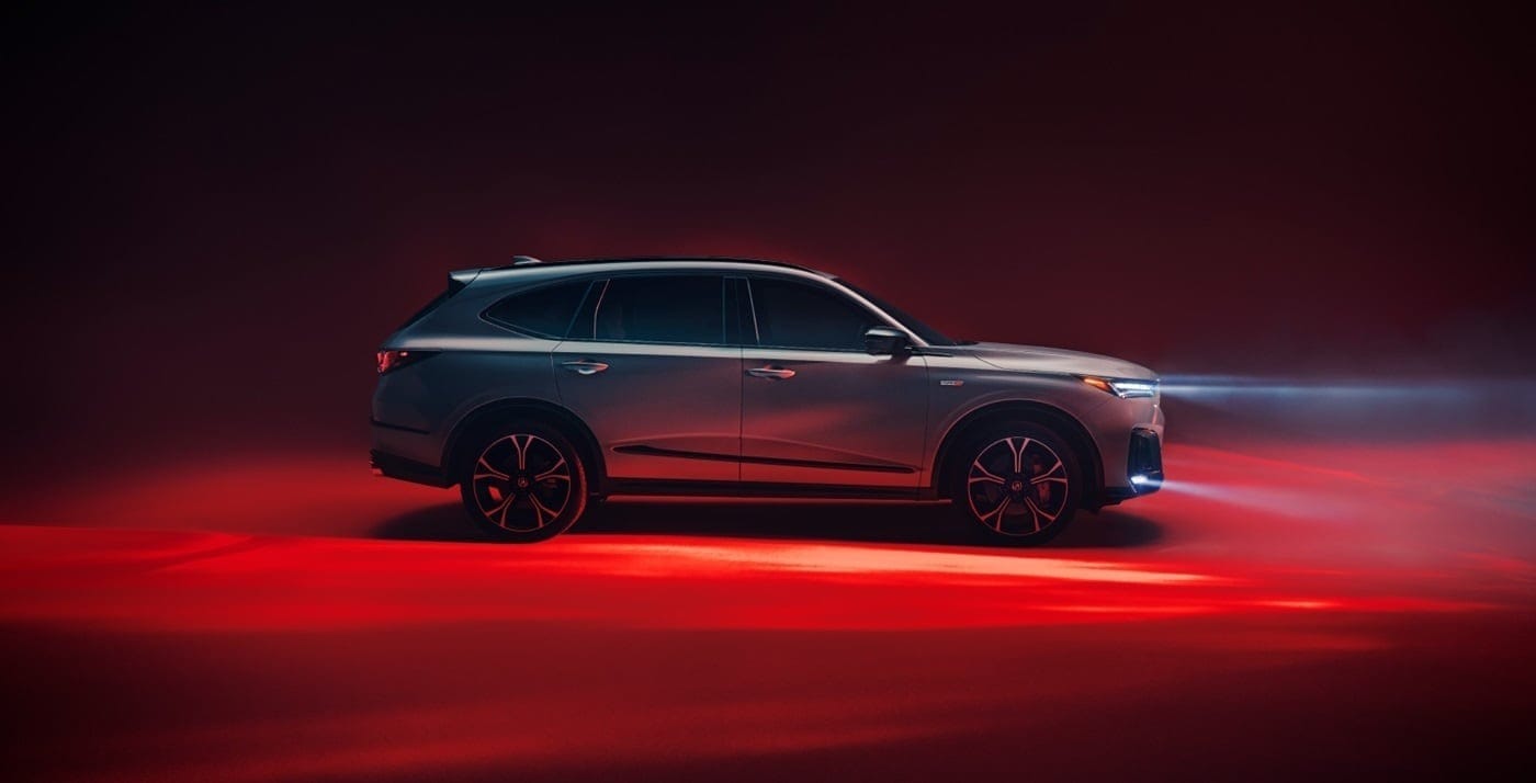 A sleek silver 2026 Acura MDX with alloy wheels, illuminated by headlights, set against a dramatic red and dark background.