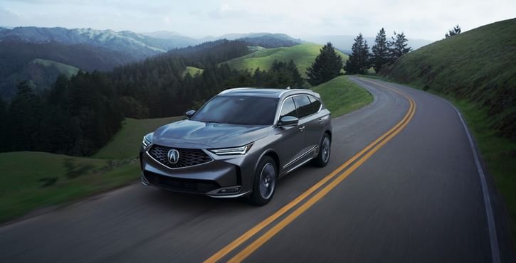 A sleek gray 2026 Acura MDX drives along a winding road, surrounded by lush green hills and distant mountains under a cloudy sky.