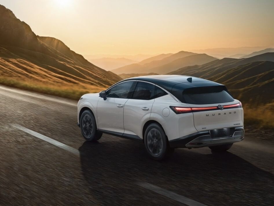 A white 2026 Nissan Murano driving on a winding road, with mountains and a sunset in the background, showcasing its sleek design.