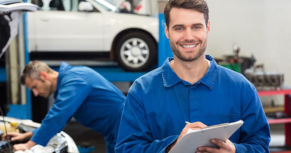 Smiling mechanic in the foreground, in the background another mechanic is repairing a car