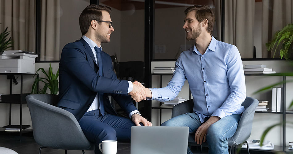 Two smiling men are sitting opposite each other and shakes hands