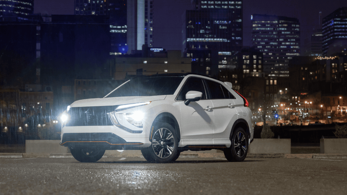 2025 Mitsubishi Eclipse Cross exterior at night, parked with headlights on against a city skyline backdrop, showcasing the SUV’s bold design and urban readiness.