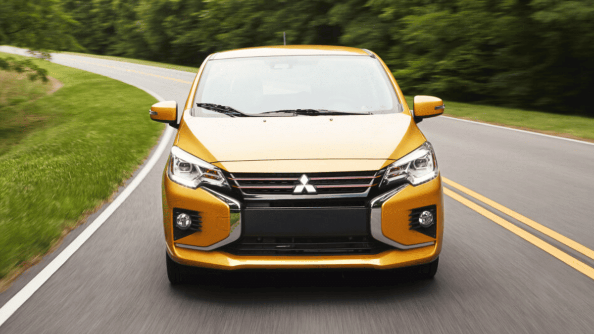 Front exterior view of the 2024 Mitsubishi Mirage, in a bold yellow color, driving down a scenic two-lane road, emphasizing its compact form and sporty styling.