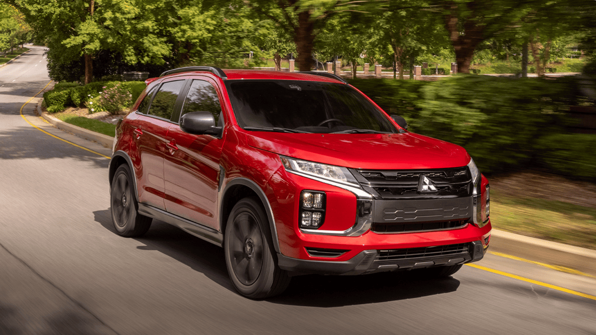 Front-side view of a red 2024 Mitsubishi Outlander Sport driving through a tree-lined road near Tampa, FL, highlighting sporty exterior styling.