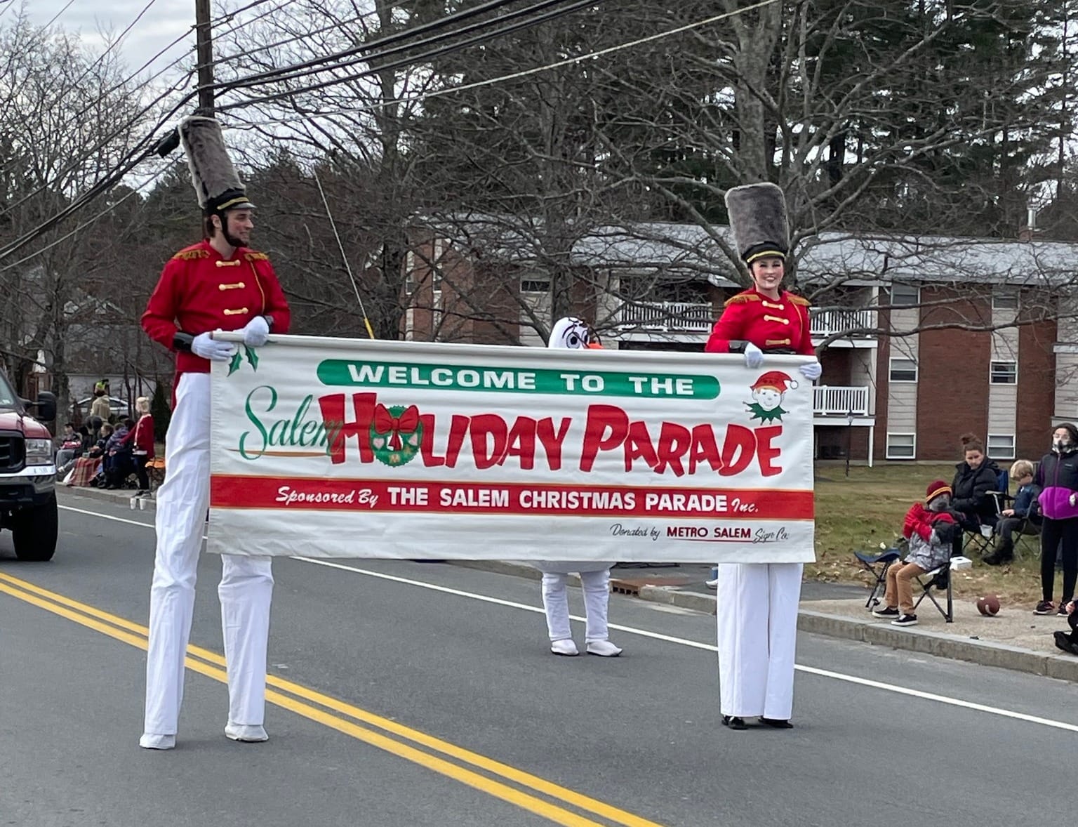 Dos zancudos vestidos como soldados de juguete sostienen un gran cartel que dice 'Bienvenidos al Desfile Navideño de Salem' mientras caminan por una calle llena de espectadores.
