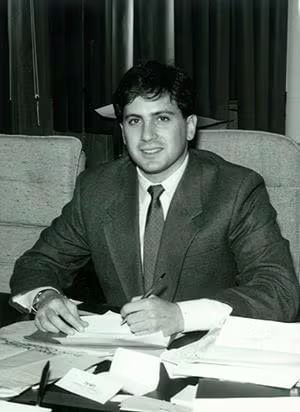 A man in a suit is seated at a desk, surrounded by papers, focused on his work.