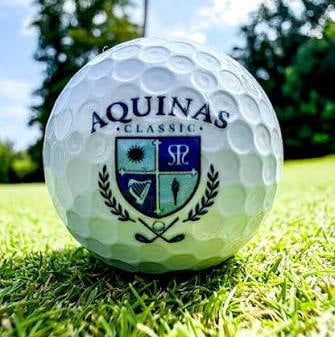 Close-up of an Aquinas Classic golf ball on bright green grass, with a flagstick and trees behind it under a sunny blue sky.