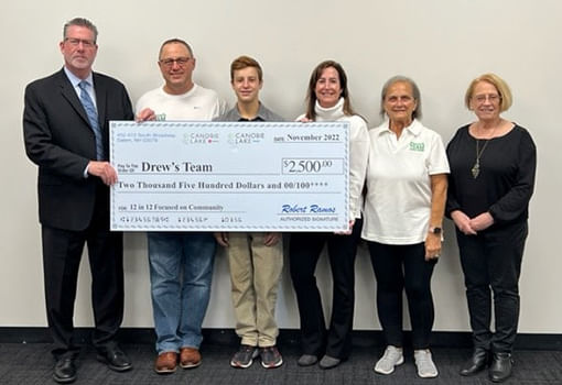 Personas se reúnen alrededor de un cheque grande, marcando una donación a Drew's Team, que ayuda a quienes son afectados por la fibrosis quística.