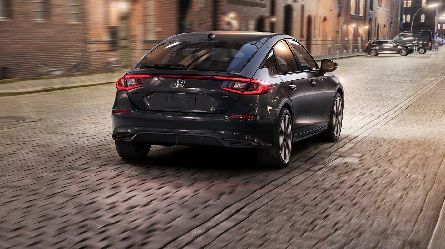 A 2026 Honda Civic Hatchback Hybrid drives on a cobblestone street lined with brick buildings. The scene conveys a modern yet historic urban feel, with evening lighting.