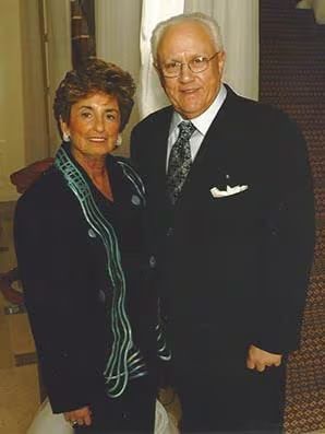 An older man and woman pose in front of a staircase, reflecting a bond of love and companionship in their expressions.