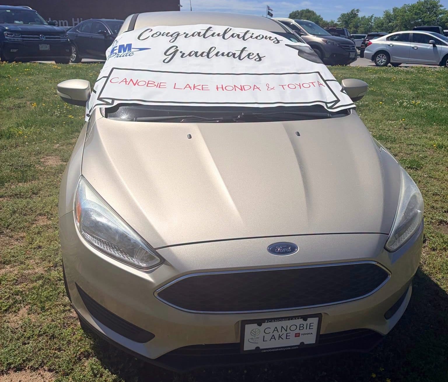 Un sedán Ford de color dorado estacionado en un terreno de césped frente a un edificio escolar de ladrillo bajo un cielo azul despejado, con una pancarta blanca extendida sobre el parabrisas delantero en la que se lee Congratulations Graduates (Felicidades graduados).