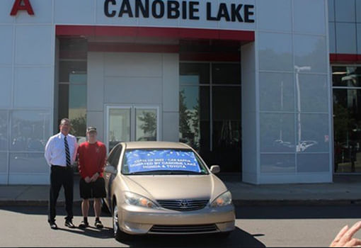 Dos hombres parados junto a un automóvil con un banner de Canobie Lake, celebrando la victoria de una rifa para un graduado de Salem High School.