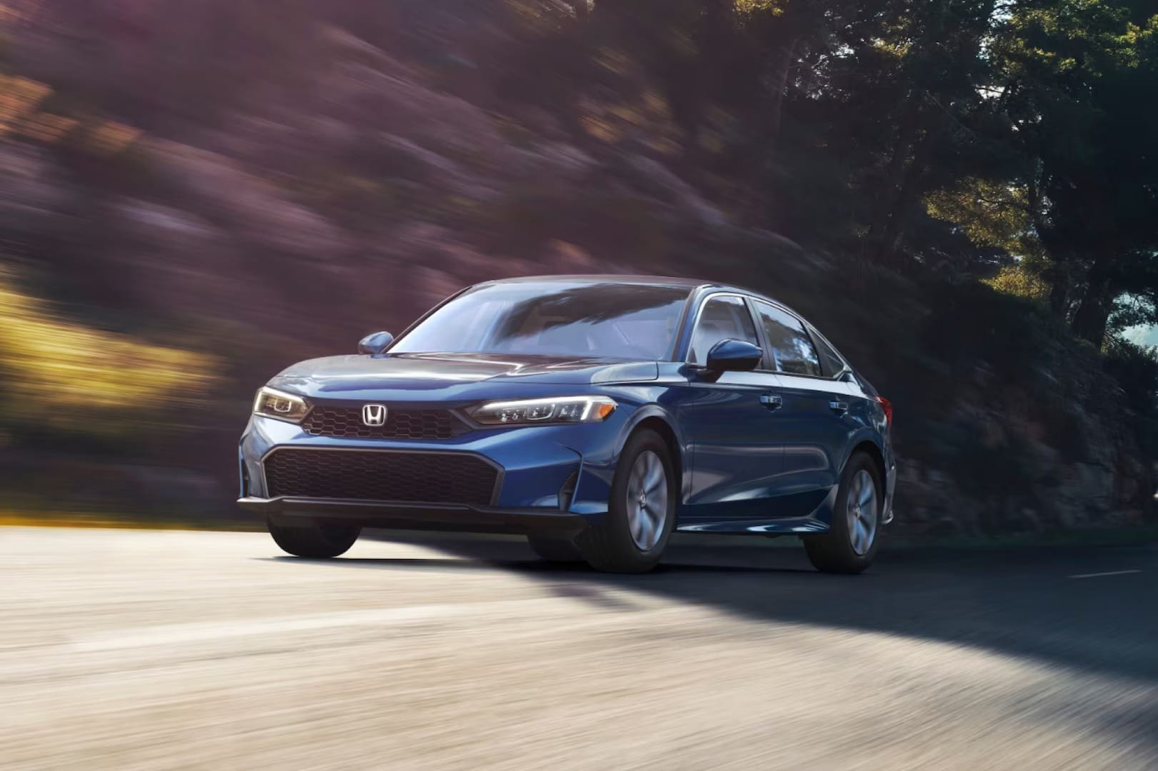 A sleek blue 2026 Honda Civic Sedan Hybrid drives swiftly on a scenic, winding road shaded by trees. The dynamic angle and blurred background convey speed and elegance.