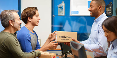 A young man and an older man at a counter receive a "Honda Genuine Parts" box from a smiling employee. Another person works at a computer nearby.
