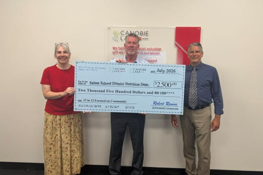 Tres personas están de pie sonriendo, sosteniendo un gran cheque por $2,500. El cheque está a nombre del Departamento de Nutrición del Distrito Escolar de Salem. El ambiente es positivo y solidario.