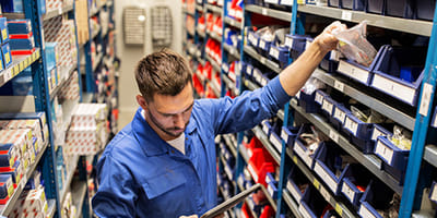 A man in a blue uniform stands in a warehouse aisle, using a tablet while selecting small parts from organized shelves. The atmosphere is focused and efficient.
