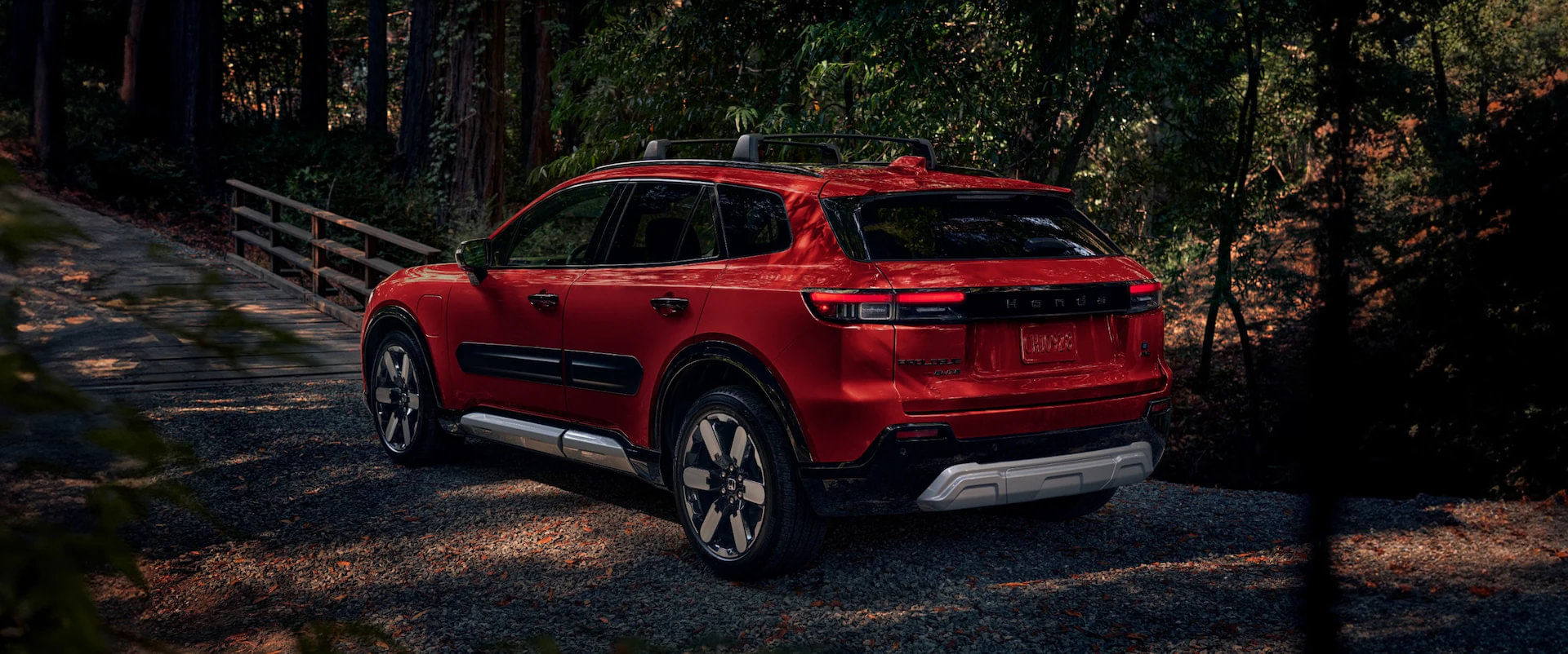 SUV Honda Prologue Elite 2025 color Rojo con portaequipajes de techo, estacionado en un camino de grava cerca de un puente peatonal de madera en un bosque bañado por el sol