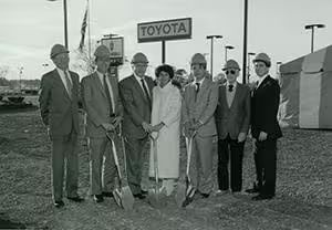 A group of people with shovels stands in front of a Toyota sign, marking the future site of a dealership.