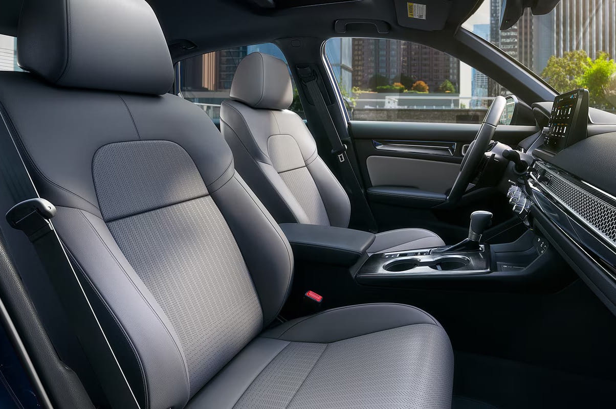 2026 Honda Civic Sedan interior with two gray leather seats, sleek dashboard, and digital display. Sunlight highlights a cityscape through the windows, creating a luxurious feel.