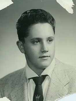 A vintage portrait of a young man with neatly styled dark hair, wearing a suit jacket, white shirt, and a tie featuring a small emblem.