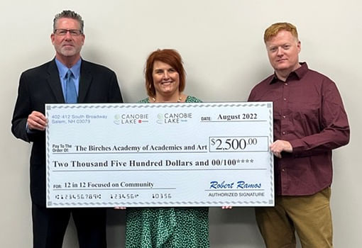 Tres personas muestran con orgullo un cheque de $2,500, apoyando la misión de The Birches Academy en educación integrada en artes.