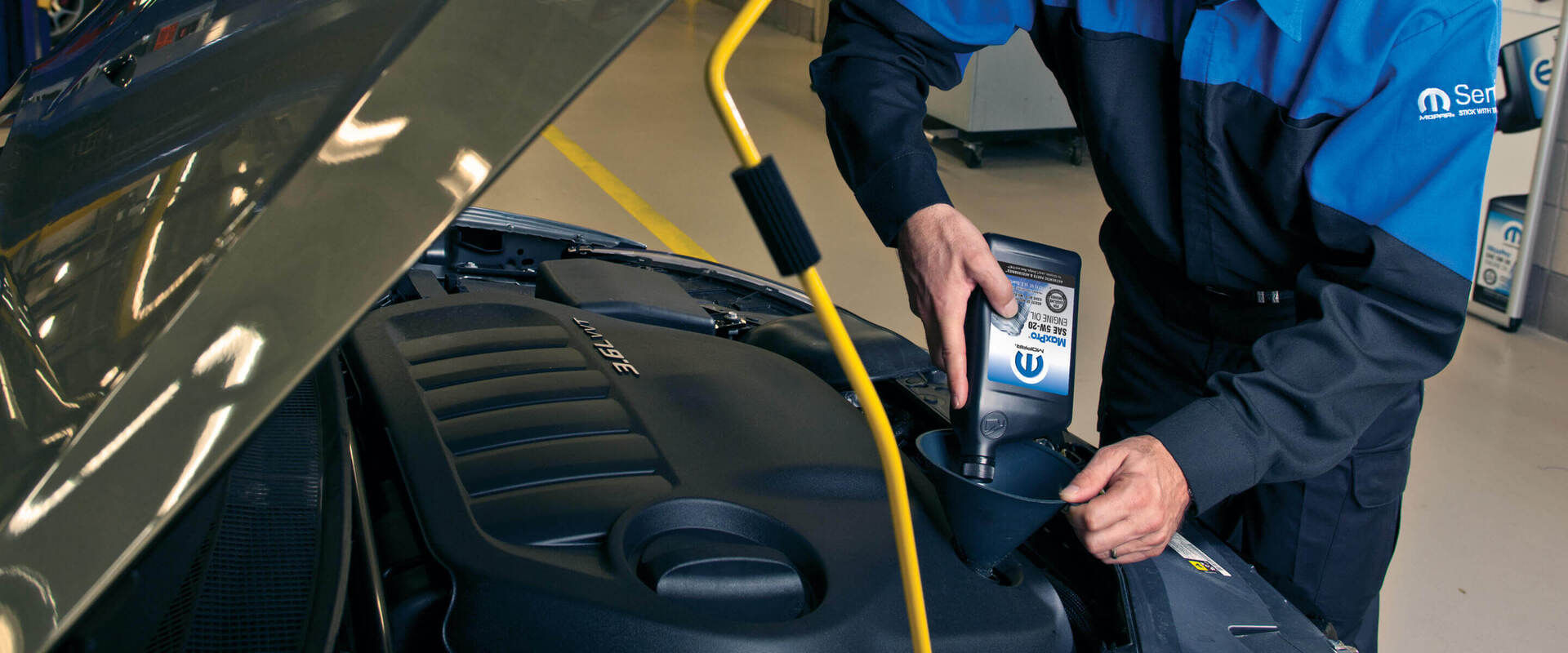A mechanic in a blue uniform adds engine oil to a car using a funnel, showing focused precision. The car hood is open, revealing the engine.