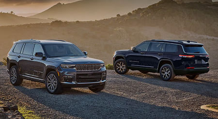 A gray 2025 Jeep Grand Cherokee Summit Reserve and a blue Jeep Grand Cherokee Limited parked opposite of each other on a gravel mountain road at sunset.
