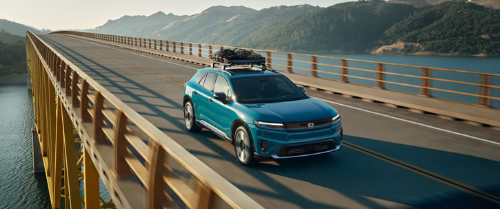 A teal 2025 Honda Prologue SUV with a roof rack drives across a sunny bridge spanning a reservoir, with rolling hills in the background.