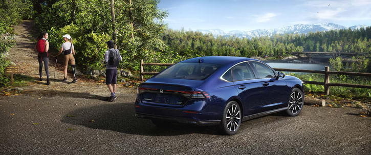 Dark blue 2025 Honda Accord, viewed from the rear passenger side, parked on asphalt near a hiking trail entrance, with a scenic background of a lake, forested hills, and mountains.