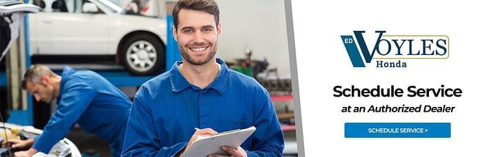 Two mechanics with one working on a car and another smiling with paper and pen