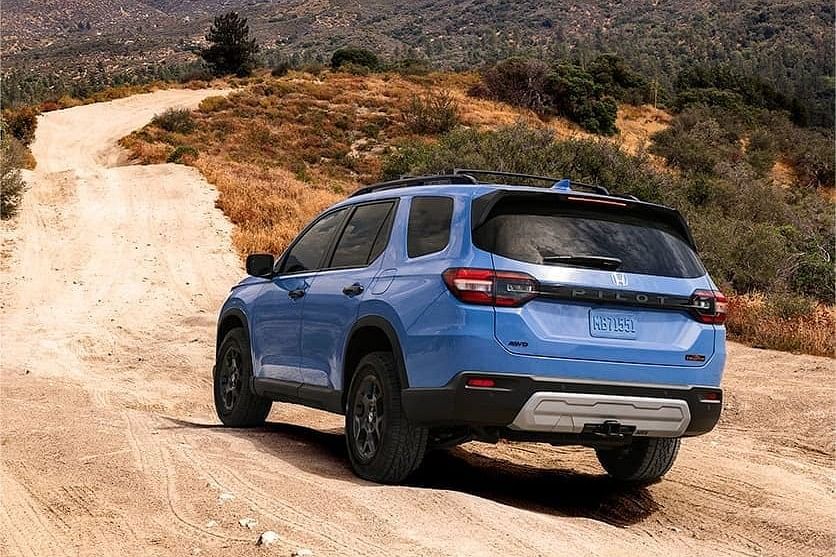A blue 2025 Honda Pilot on a dirt road, surrounded by hills and greenery, ready for adventure in a rugged landscape.