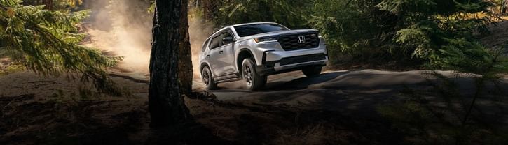 A silver 2025 Honda Pilot navigates a dusty forest path, surrounded by tall trees and sunlight filtering through the leaves.