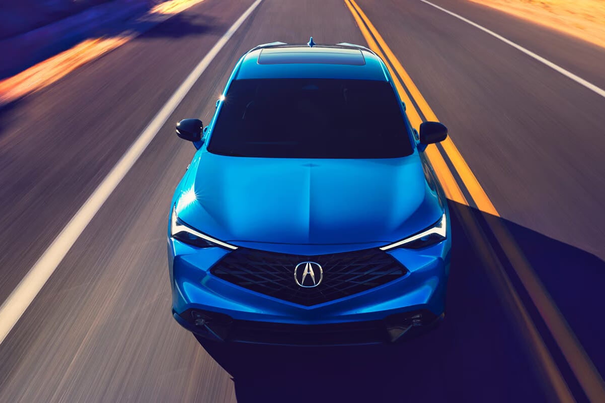 A blue 2026 Acura ADX is driving on an empty road with double yellow lines. The scene is captured from above, highlighting the vehicle's sleek design and dynamic setting.