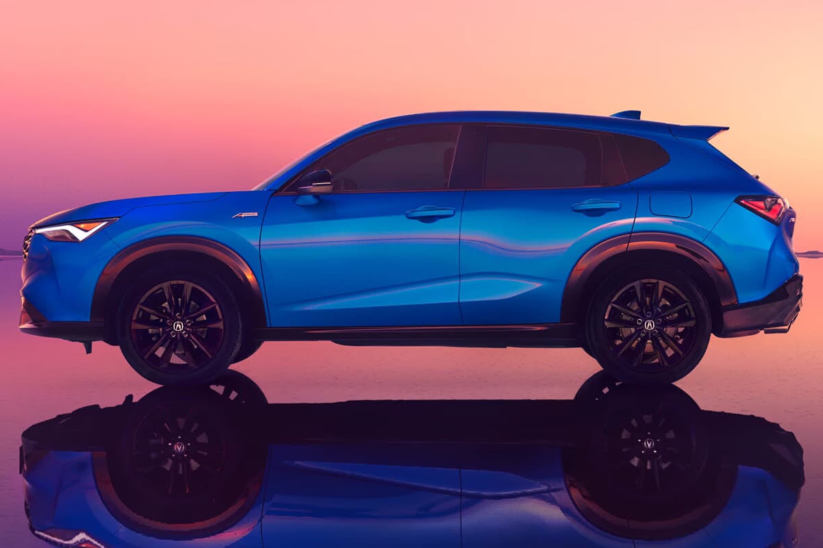 A blue 2026 Acura ADX is parked on a reflective surface against a vibrant pink and orange sunset. The sleek design and warm lighting create a modern, serene atmosphere.