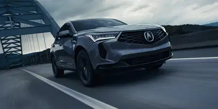 A dark gray 2026 Acura RDX driving dynamically on a bridge under an overcast sky, highlighting its diamond pentagon grille and sharp LED headlights.