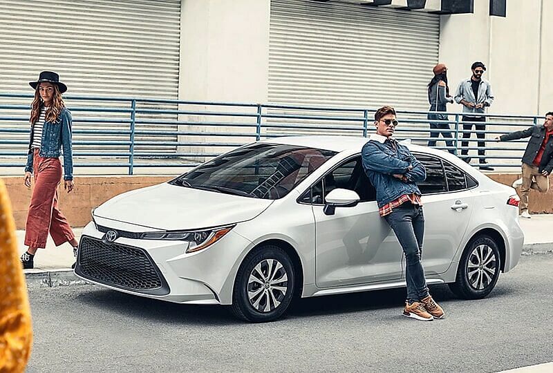 Young guy leaned up against a parked 2022 Toyota Corolla Hybrid while onlookers pass by