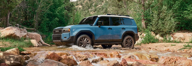 A blue 2026 Toyota Land Cruiser navigating through a rocky creek surrounded by lush greenery and trees, showcasing its rugged off-road capability.