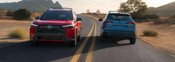 A red and a blue Toyota Corolla Cross on a road in a scenic, rocky, desert landscape.