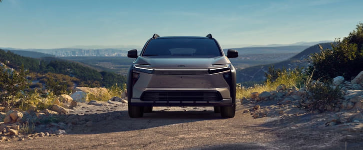 Front view of a sleek gray SUV on a rocky dirt road with mountains and cityscape in the background under clear skies.