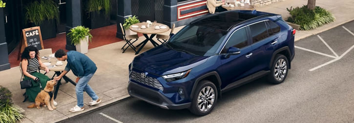 A dark blue Toyota RAV4 SUV parked on a street next to an outdoor cafe seating area, where a couple interacts with a dog.