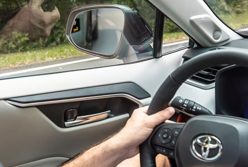 Interior view of a car from the driver's perspective, showing a hand on the steering wheel and the side-view mirror with a blind-spot monitoring icon illuminated.