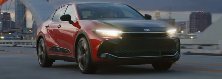 Toyota Crown sedan driving across a city bridge at dusk with skyline in the background.