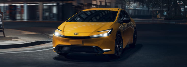A front view of a yellow 2026 Toyota Prius driving on a city street at night.